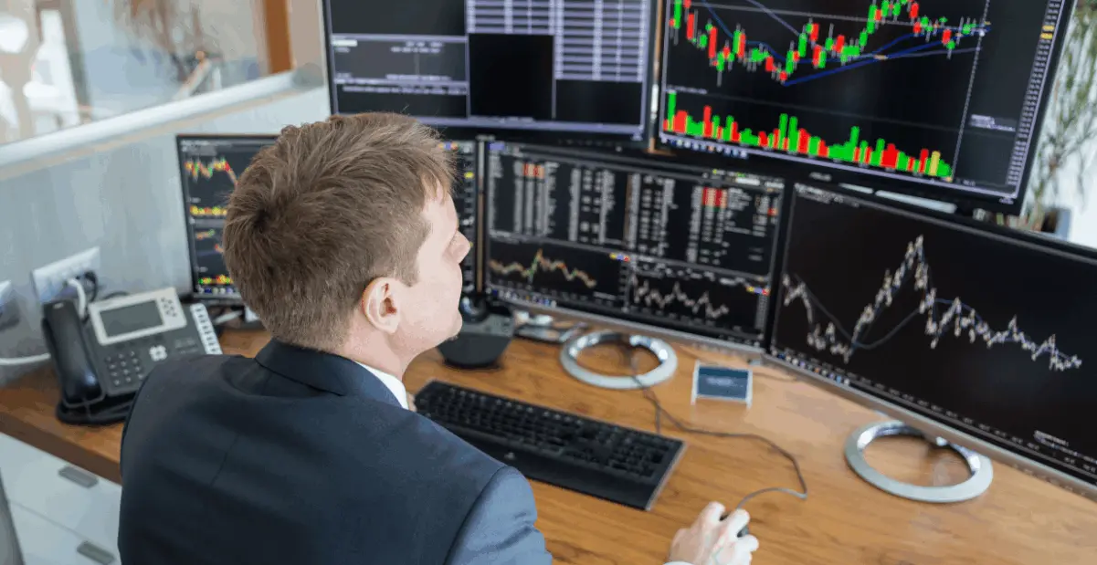 A trader sits at a desk with multiple computer monitors displaying various stock charts and financial data.