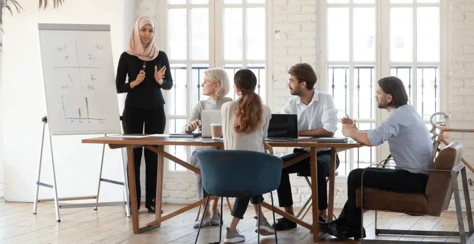 A woman presenting data to colleagues seated at a conference table, gesturing towards a flip chart with graphs and charts